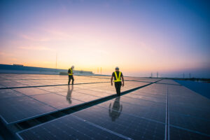 Two workers in high visibility outerwear walk across solar panels with a sunset in the background.
